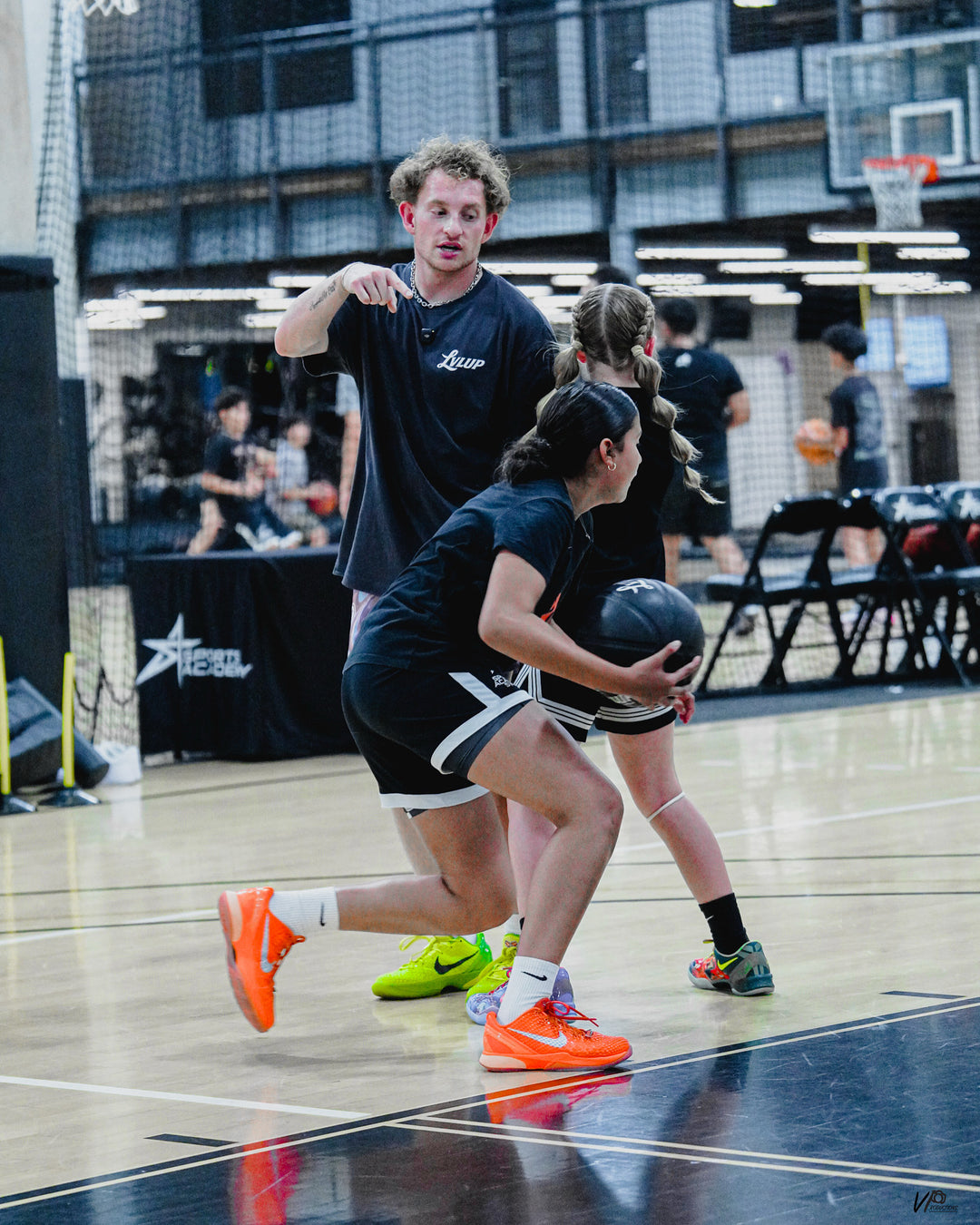 Basketball trainer coaching two young athletes during a drill, as one player drives forward holding a black LVLUP Handle heavy ball on an indoor court.