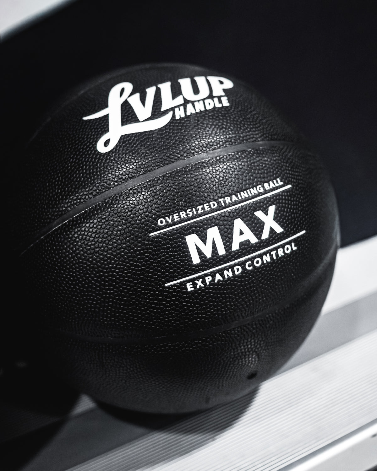 A close-up of a black textured training ball with white printed text reading 'LvLup Handle' and 'MAX Expand Control', resting on a metallic surface against a dark background.