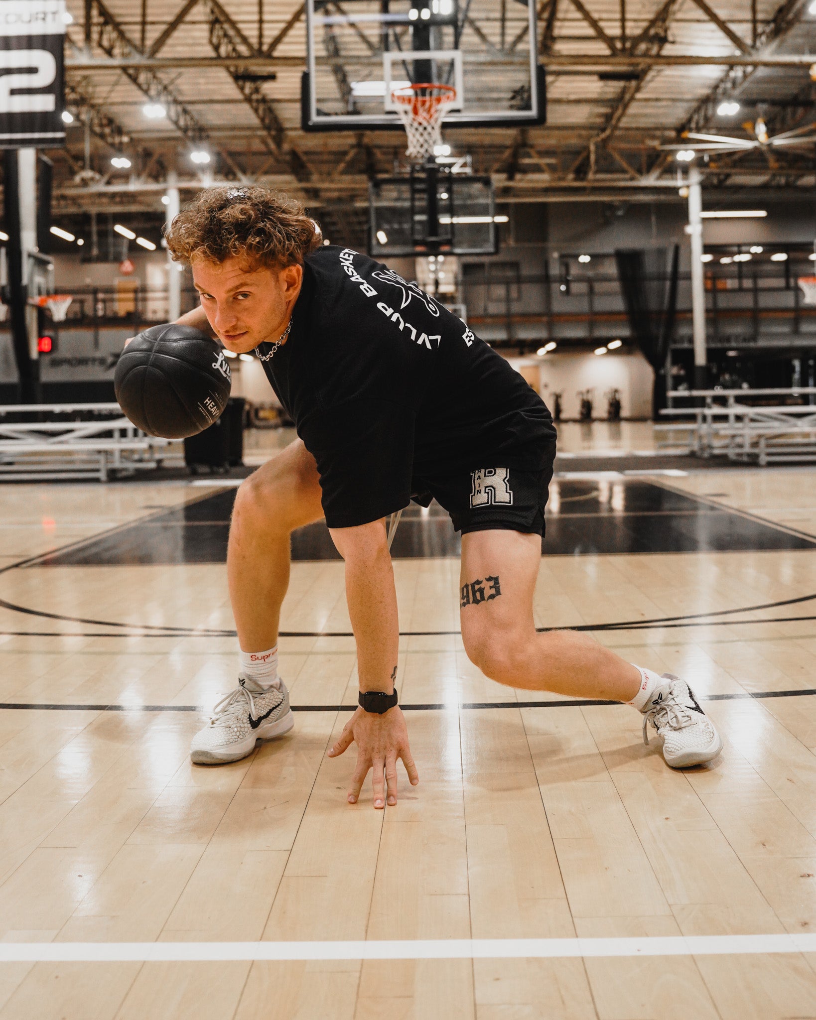 Basketball trainer in a low crossover stance dribbling a black LVLUP Handle heavy ball on an indoor court, wearing black training gear and white sneakers.