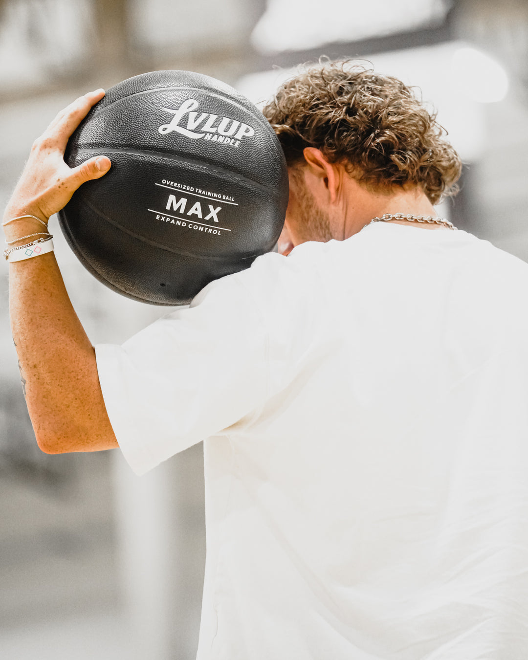 A man in a white t-shirt holding a black Lulup Max basketball close to his face, shown from behind in a bright, outdoor setting.