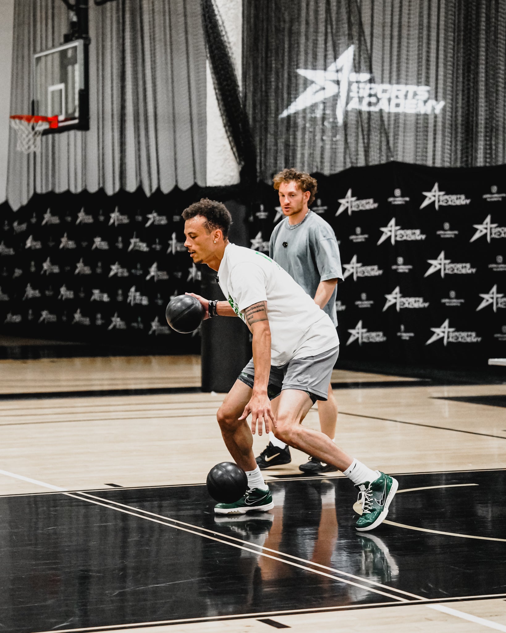 A man in a white t-shirt and gray shorts dribbling a black basketball on an indoor basketball court, with another man in a gray shirt standing behind him. The background features a black banner with white logos and a basketball hoop.