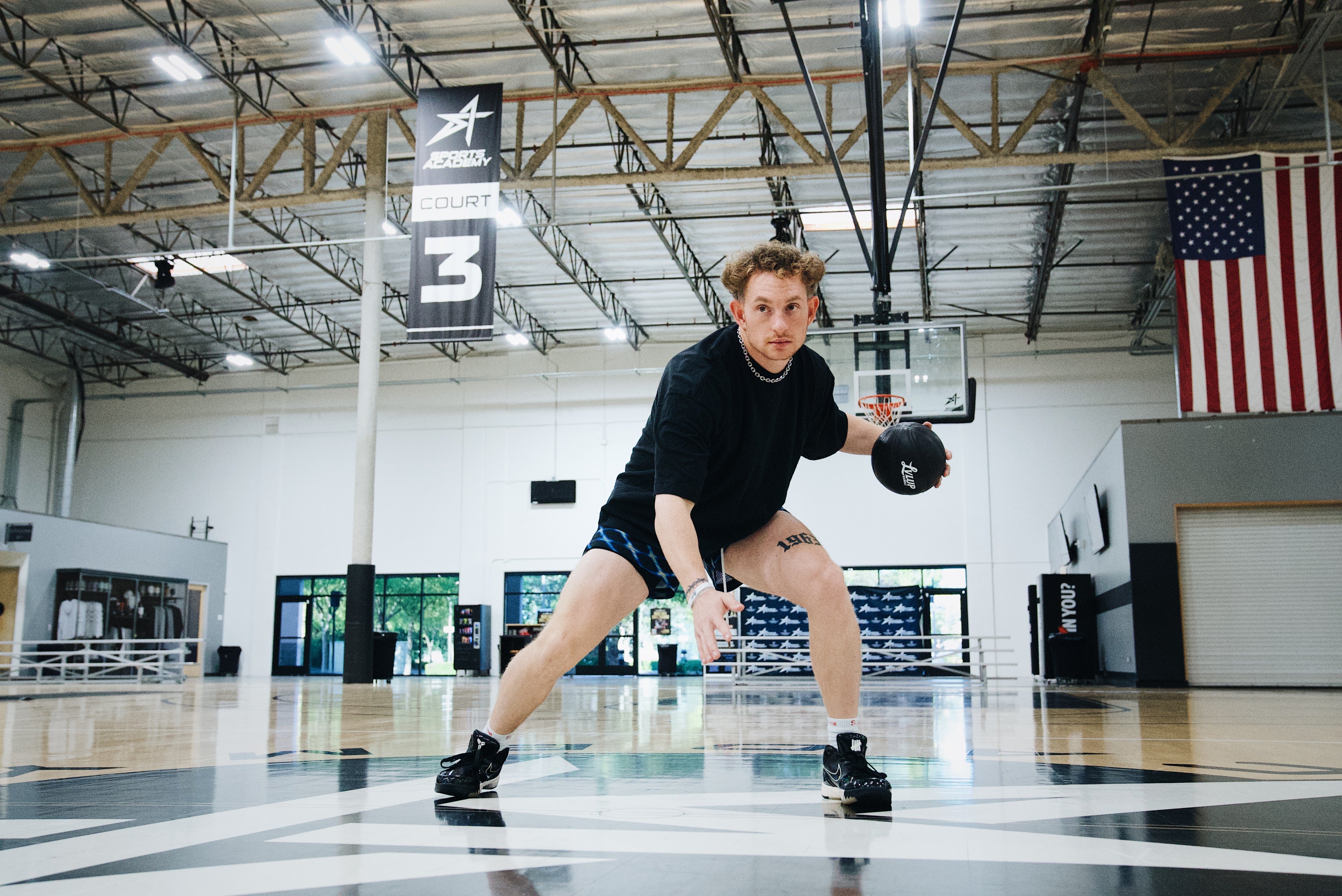 Basketball trainer in a wide stance dribbling a black LVLUP Handle heavy ball on an indoor court, locked in on the drill with the Sports Academy Court 3 banner overhead.