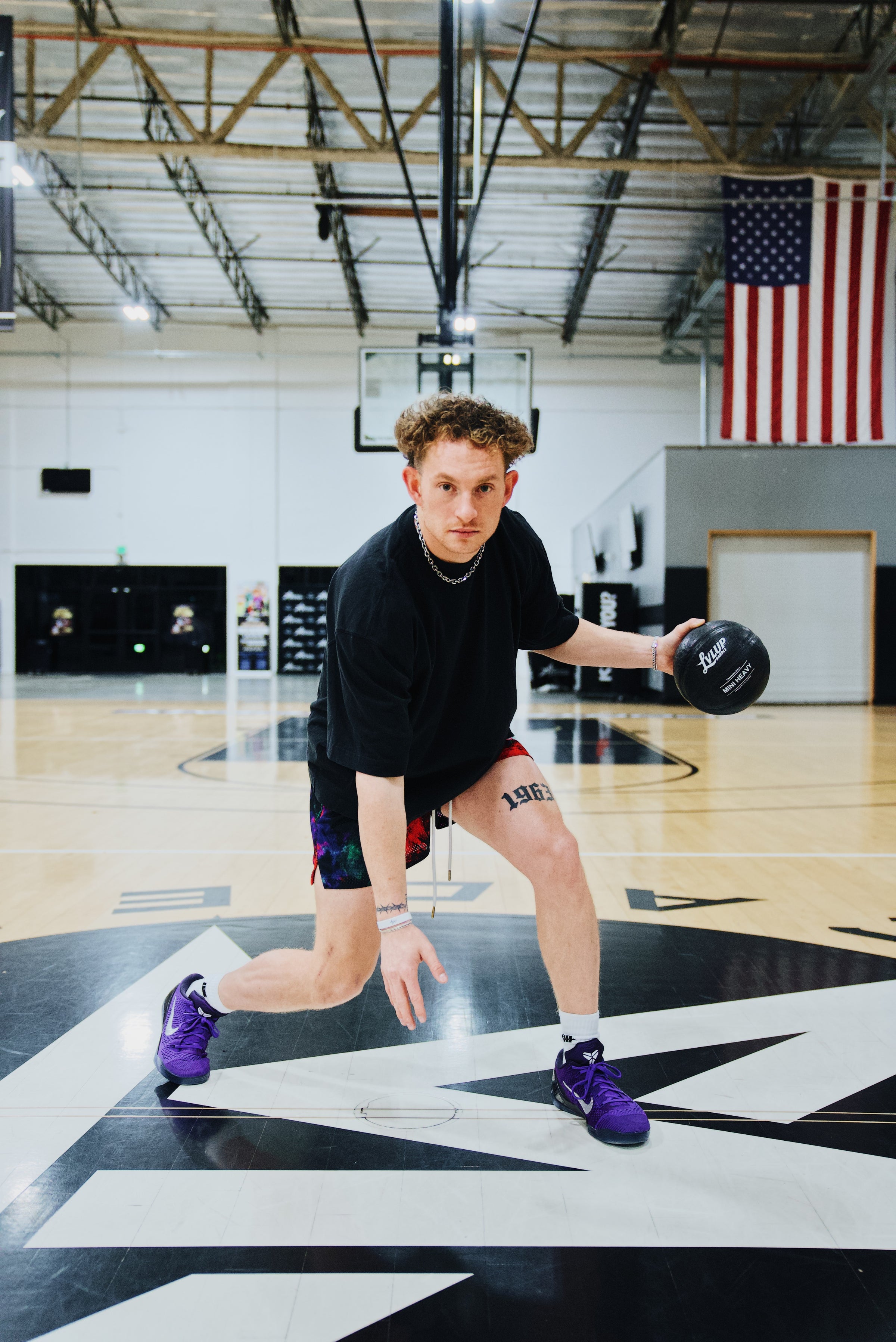 Basketball trainer performing a low dribble move on an indoor court while holding a black LVLUP Handle heavy ball, wearing a black shirt, colorful shorts, and purple basketball shoes.