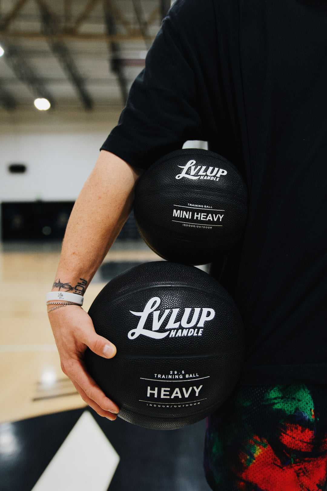 Close-up of a person holding two black LVLUP Handle weighted basketballs, the Mini Heavy under their arm and the Heavy ball in their hand, showing both logos clearly on an indoor court.