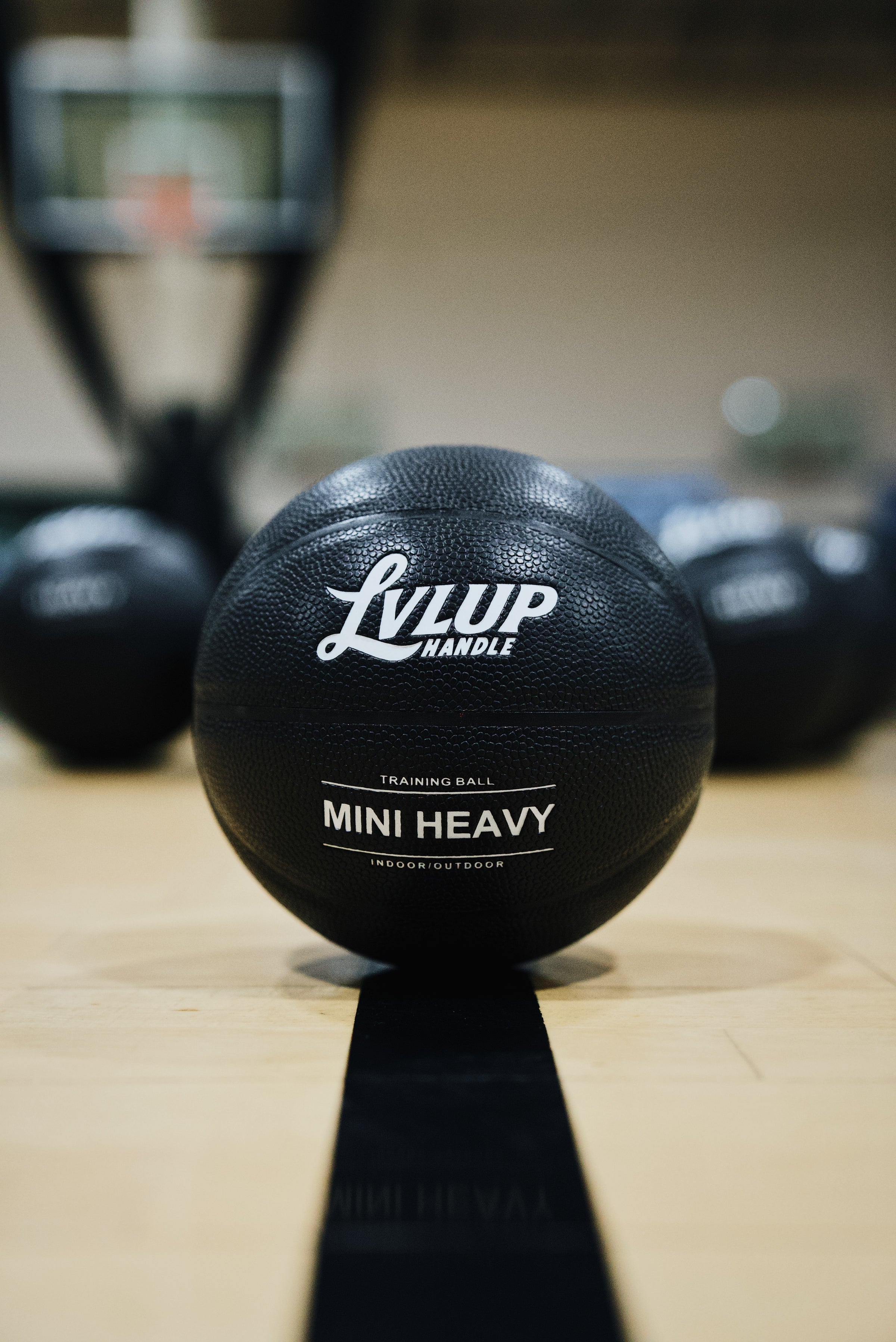 Close-up of a black LVLUP Handle Mini Heavy training ball on a basketball court, with additional weighted balls blurred in the background.