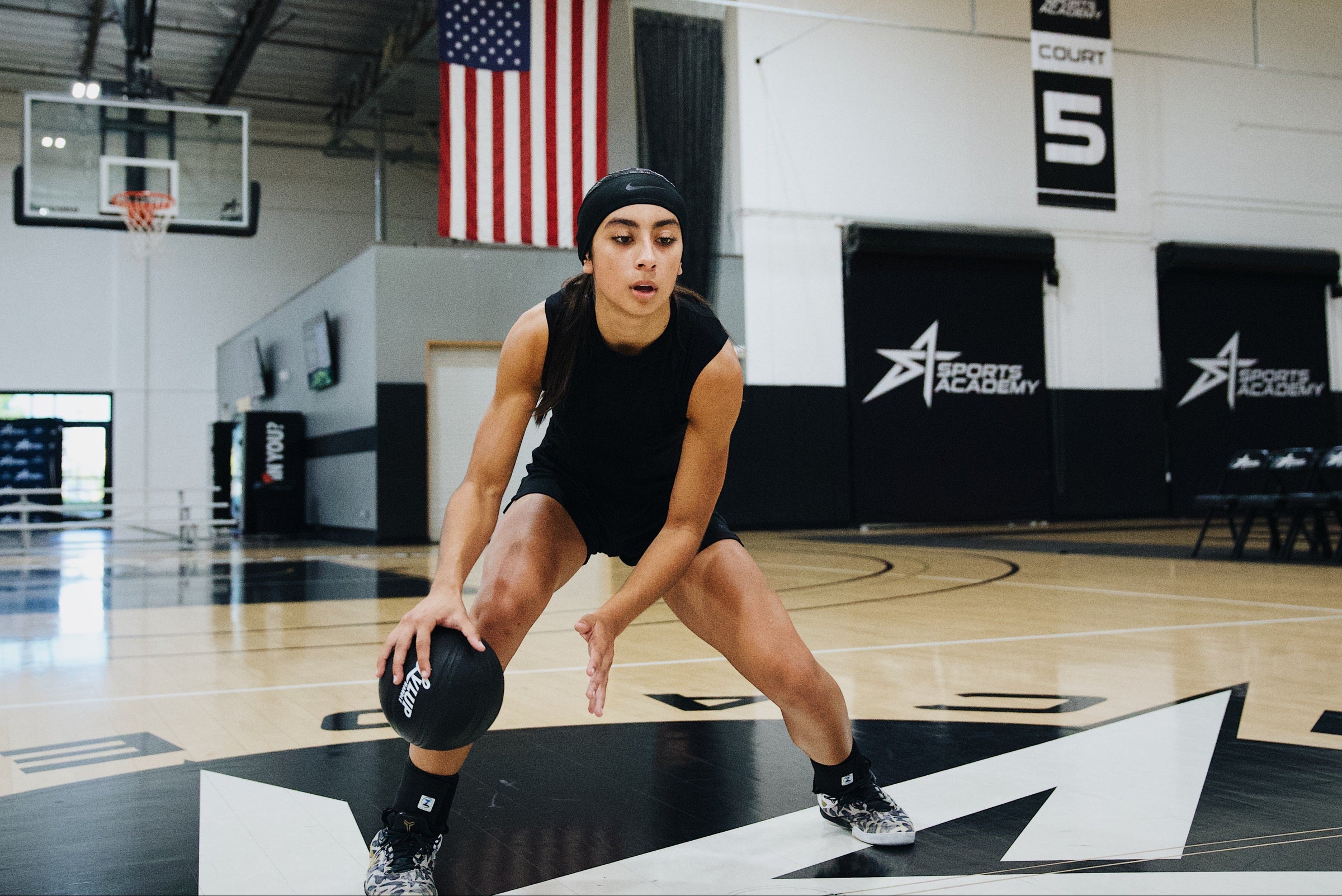 Female basketball athlete in a low stance dribbling a black LVLUP Handle heavy ball on an indoor court, wearing all-black training gear and staying locked in on the drill.