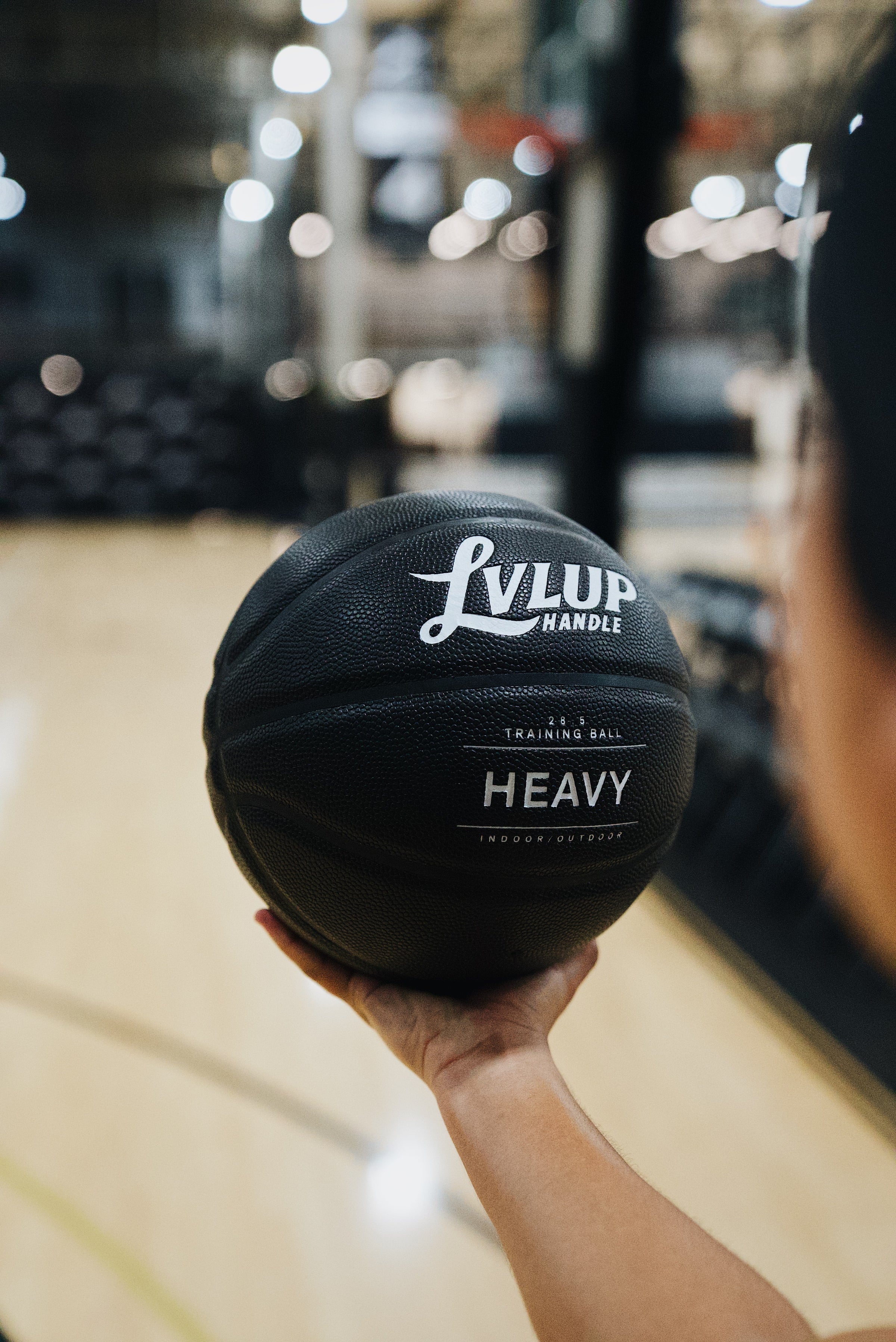 Close-up of a person holding a black LVLUP Handle Heavy training basketball in one hand, with the logo and textured surface in sharp focus against a blurred indoor court.