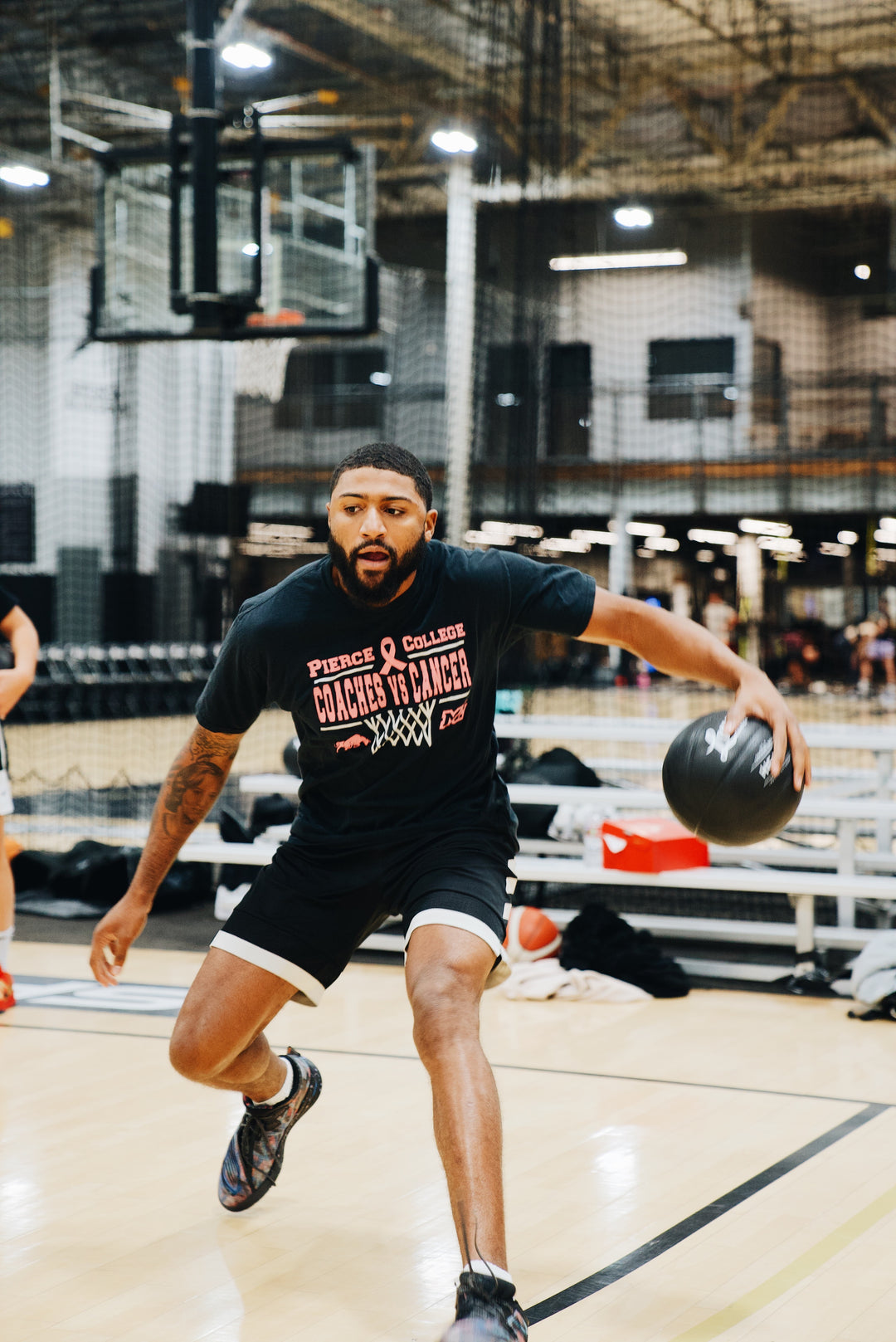 Basketball player driving forward while dribbling a black LVLUP Handle heavy ball on an indoor court, wearing a Pierce College Coaches vs Cancer shirt and black training gear.