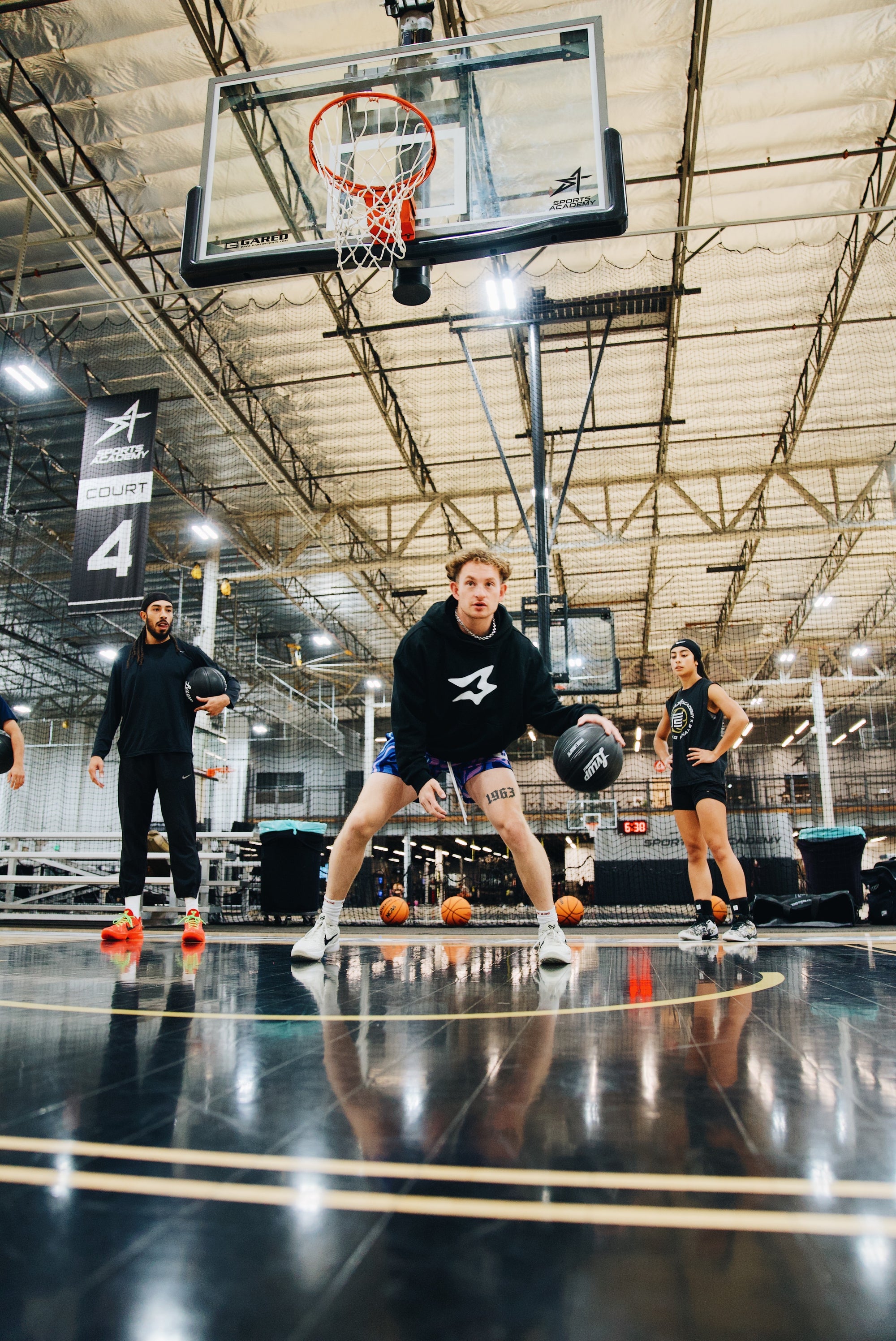 Group of basketball players training on an indoor court, with the athlete in front dribbling a black LVLUP Handle heavy ball in a low stance while others watch in the background under a Sports Academy Court 4 banner.