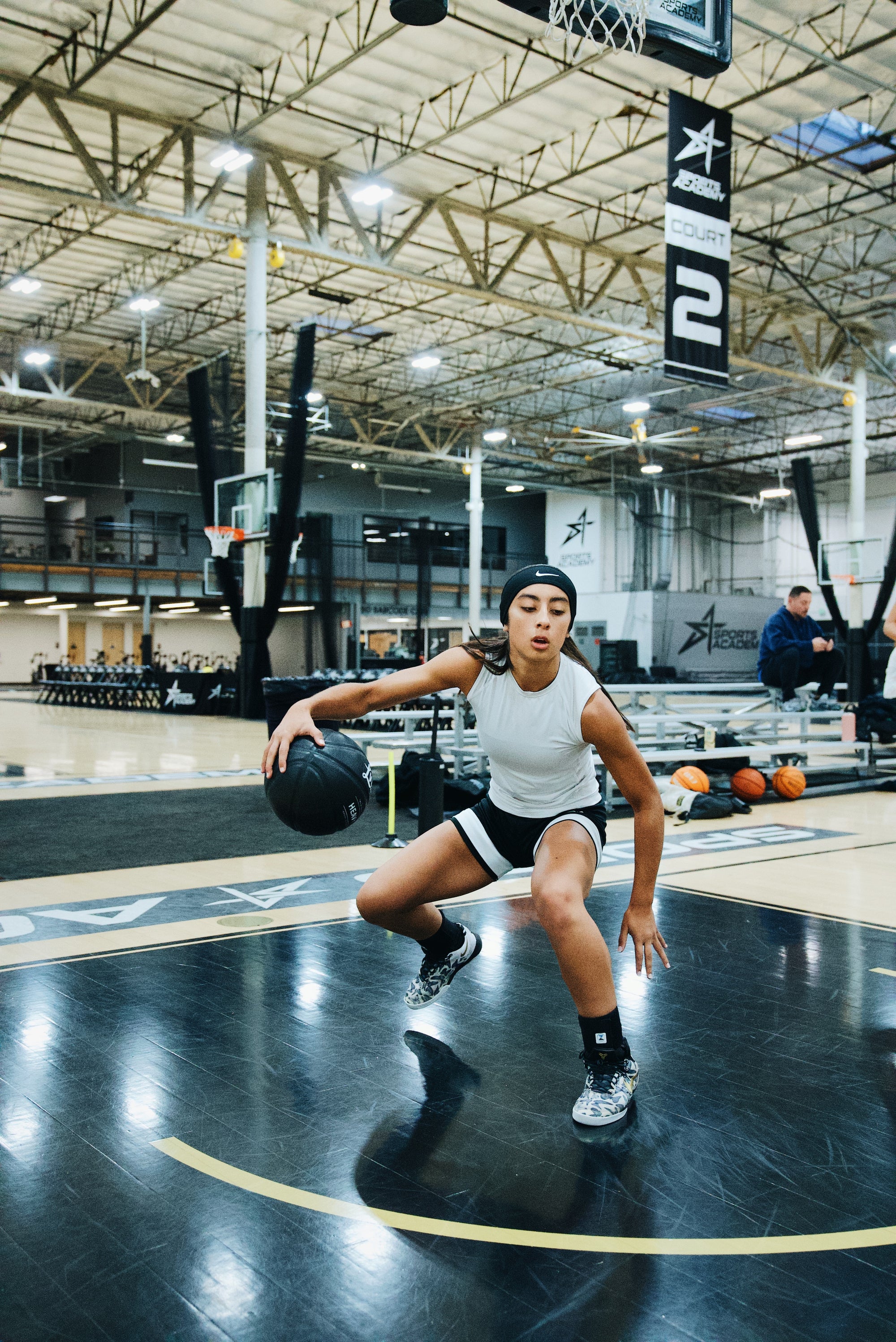 Female basketball athlete in a low stance dribbling a black LVLUP Handle heavy ball on an indoor court, wearing a white top, black shorts, and a headband.
