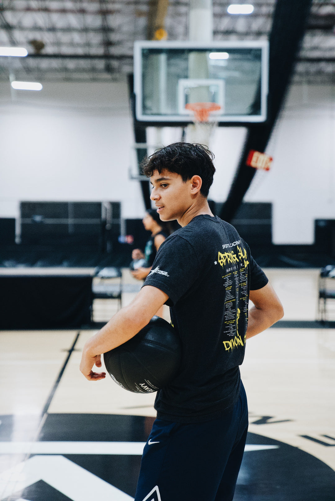 Young basketball player holding a black LVLUP Handle heavy ball at their side on an indoor court, looking toward the action while wearing a dark graphic t-shirt and navy shorts.