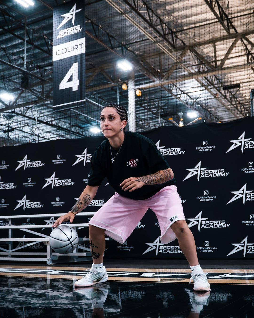A man wearing a black t-shirt, light pink shorts, and white sneakers dribbles a basketball on an indoor court, with a Sports Academy backdrop behind him.