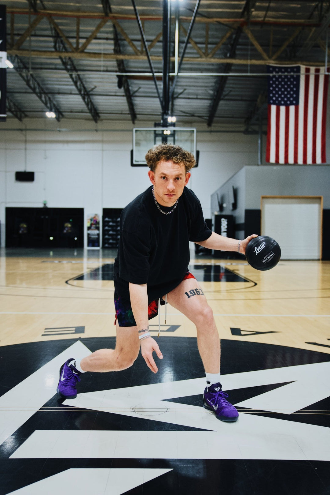 Basketball trainer in a low dribble stance on an indoor court, controlling a black LVLUP Handle heavy ball while wearing a black shirt, colorful shorts, and purple sneakers.