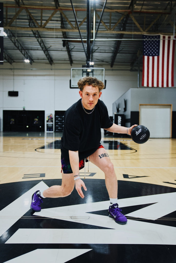 Basketball trainer in a low dribble stance on an indoor court, controlling a black LVLUP Handle heavy ball while wearing a black shirt, colorful shorts, and purple sneakers.